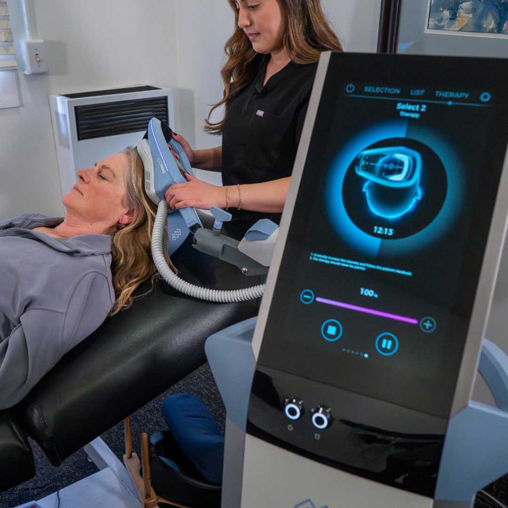 Clinician administering an Exomind neurostimulation treatment to a patient reclining on a treatment table, with the Exomind control system visible in the foreground.