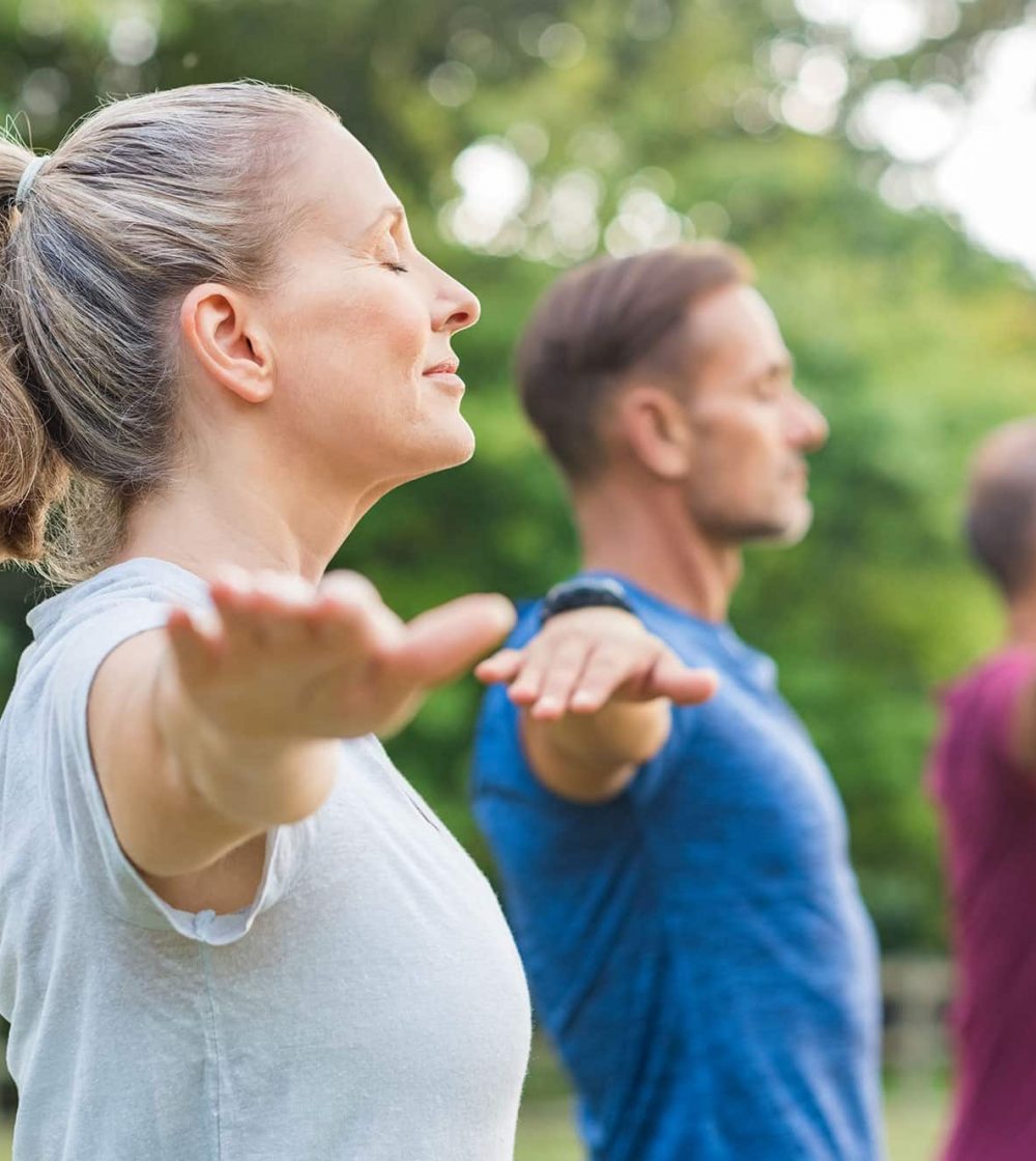 Three adults practicing mindful movement and deep breathing outdoors in a green park setting, with arms extended and eyes closed.